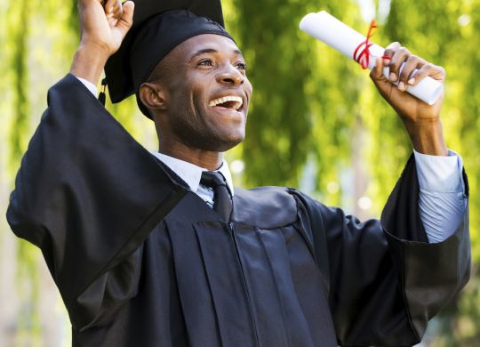 Finally graduated! Happy young African man in graduation gowns holding diploma and rising arms up