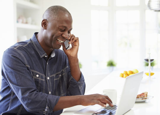 Man Using Laptop And Talking On Phone In Kitchen At Home