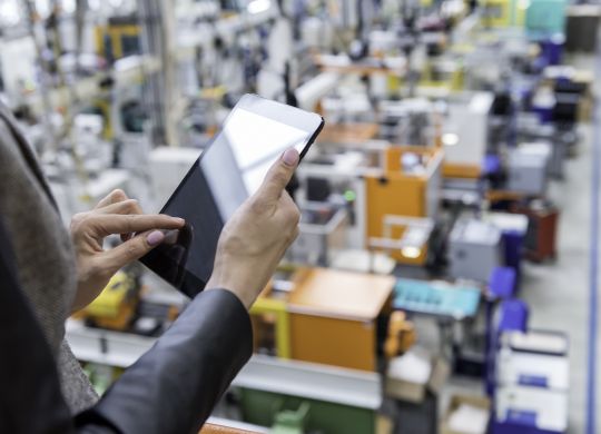 Horizontal color image of businesswoman - unrecognizable person - working with digital tablet in large futuristic factory. Woman standing on top of a balcony, holding touchpad and checking inventory of a manufacturing company on touchscreen tablet. Focus on businesswoman's hands holding black tablet, futuristic machines in background.